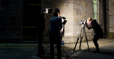 A female journalist at a camera filming an interview subject who is obscured by the lighting techs.