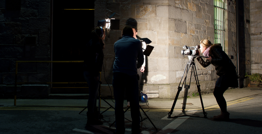 A female journalist at a camera filming an interview subject who is obscured by the lighting techs.