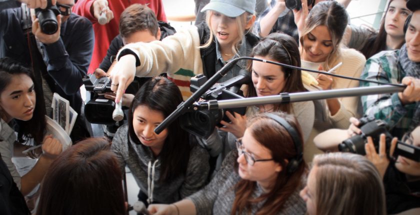 Mostly female journalists scrum interviewing someone. Only the interviewees hair can be seen from behind.