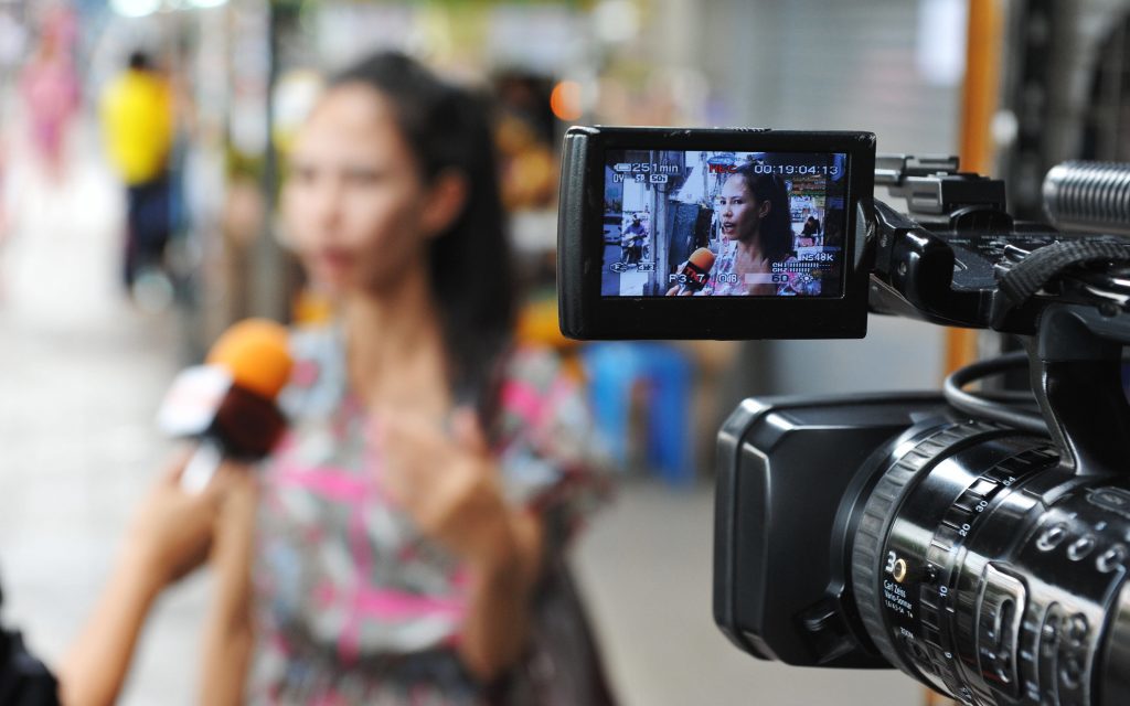A woman is being interviewed out of focus in the background. A camera is set up and filming the woman. There is an in focus shot of her in the view finder.