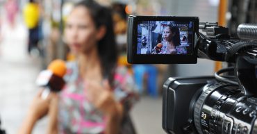 A woman is being interviewed out of focus in the background. A camera is set up and filming the woman. There is an in focus shot of her in the view finder.