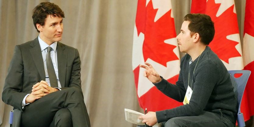 Justin Trudeau being interviewed by a journalists with Canadian flags in the background.